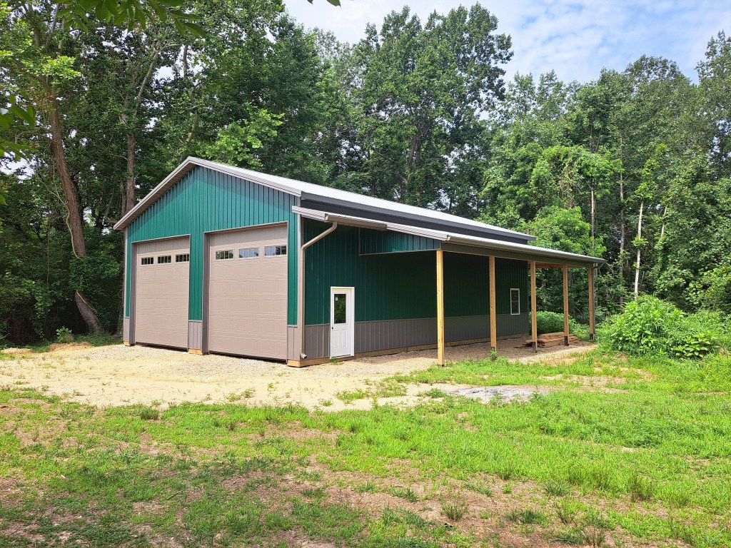 A large metal-sided barn building sits in a clearing among trees, featuring a green roof and walls. It has a gravel founda...