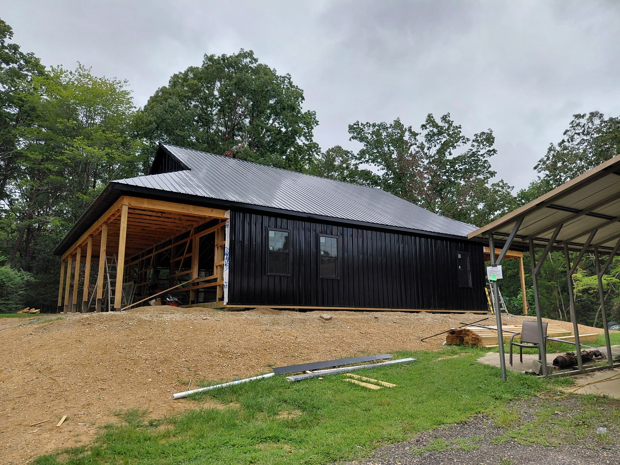 The image depicts the back of a house under construction, situated in a wooded area. The house is black with a gray roof a...